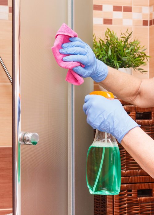 Hand of senior woman in protective gloves cleaning glass shower using microfiber cloth and detergent, concept of household duties